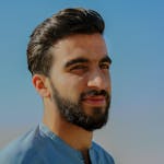 A close-up outdoor portrait of a young man with facial hair, exuding confidence under a clear sky.