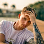 A thoughtful young man with tattoos, leaning outdoors on a sunny day.