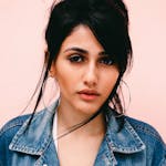 Stylish woman with dark hair posing in denim jacket against a pastel pink wall.